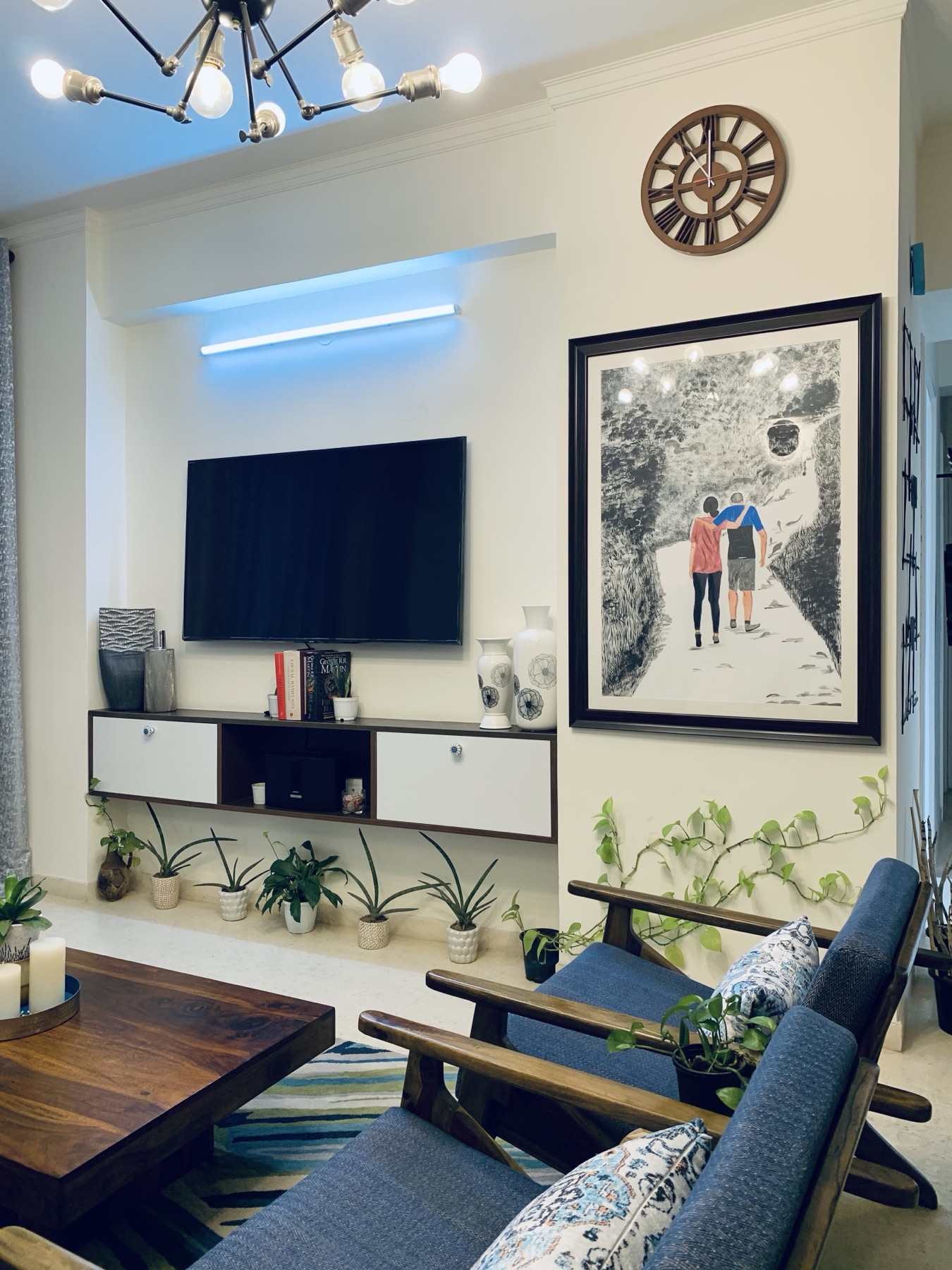 A living room wall with a television above a floating white and walnut console, a row of potted plants along the floor, and a framed drawing beside it.