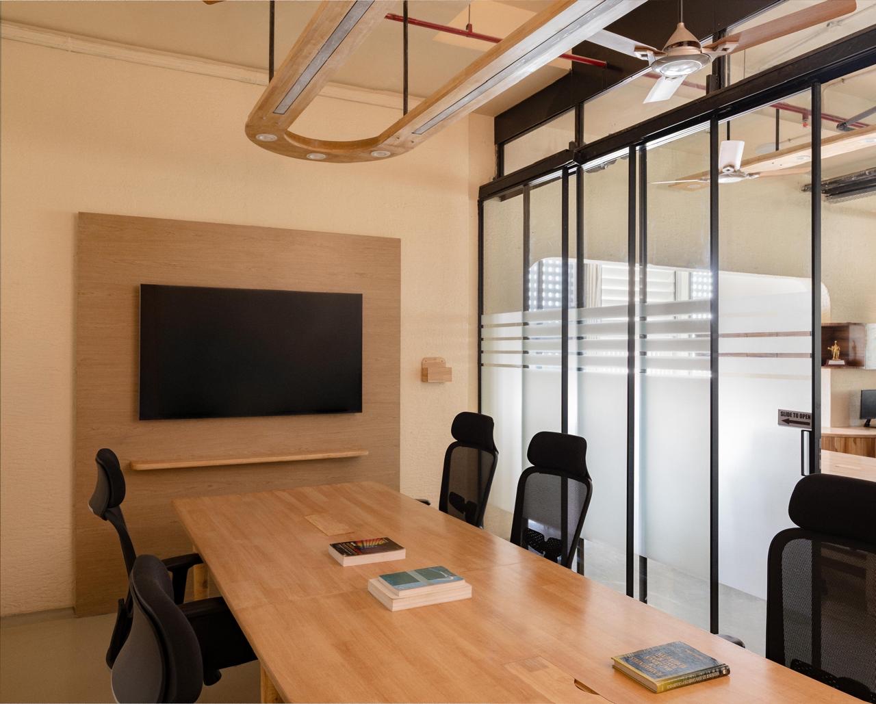 A meeting room with a long light-wood table, black mesh chairs, a screen mounted on a wood panel, a timber light fitting overhead, and sliding glass doors with frosted bands.