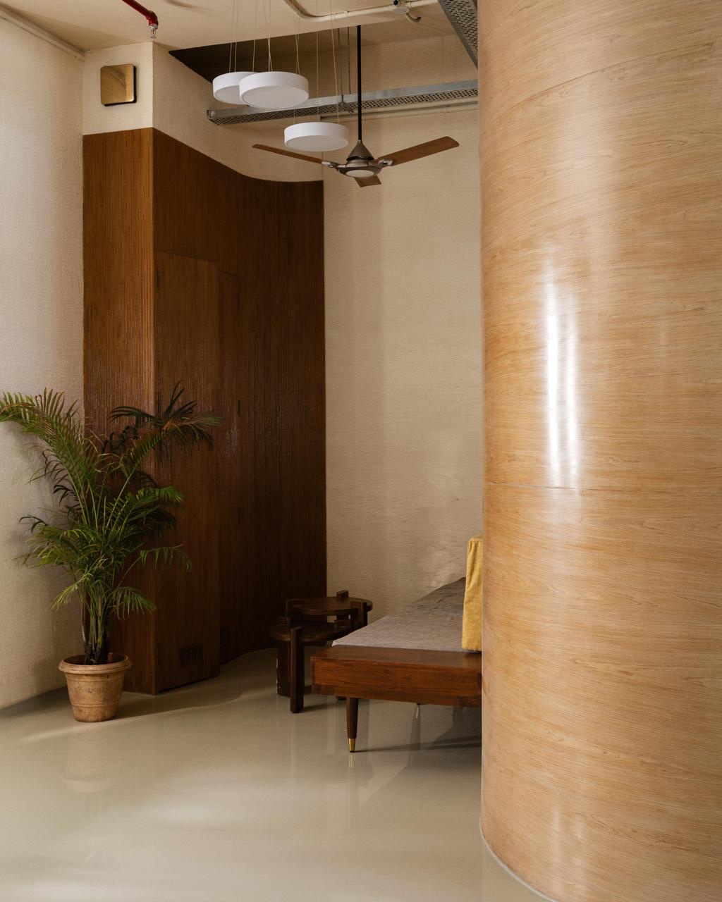 A curved veneer wall in the foreground. Beyond it are a bench, a potted palm, a fluted timber wall, and three round pendant lights.