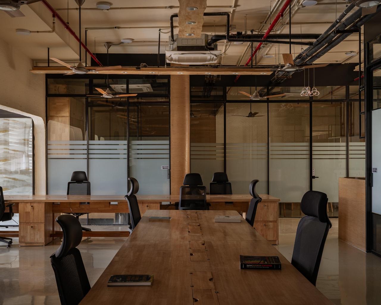 A long solid-wood table with black mesh chairs on an open office floor, with glass-fronted rooms behind it and pipes and ceiling fans left exposed overhead.
