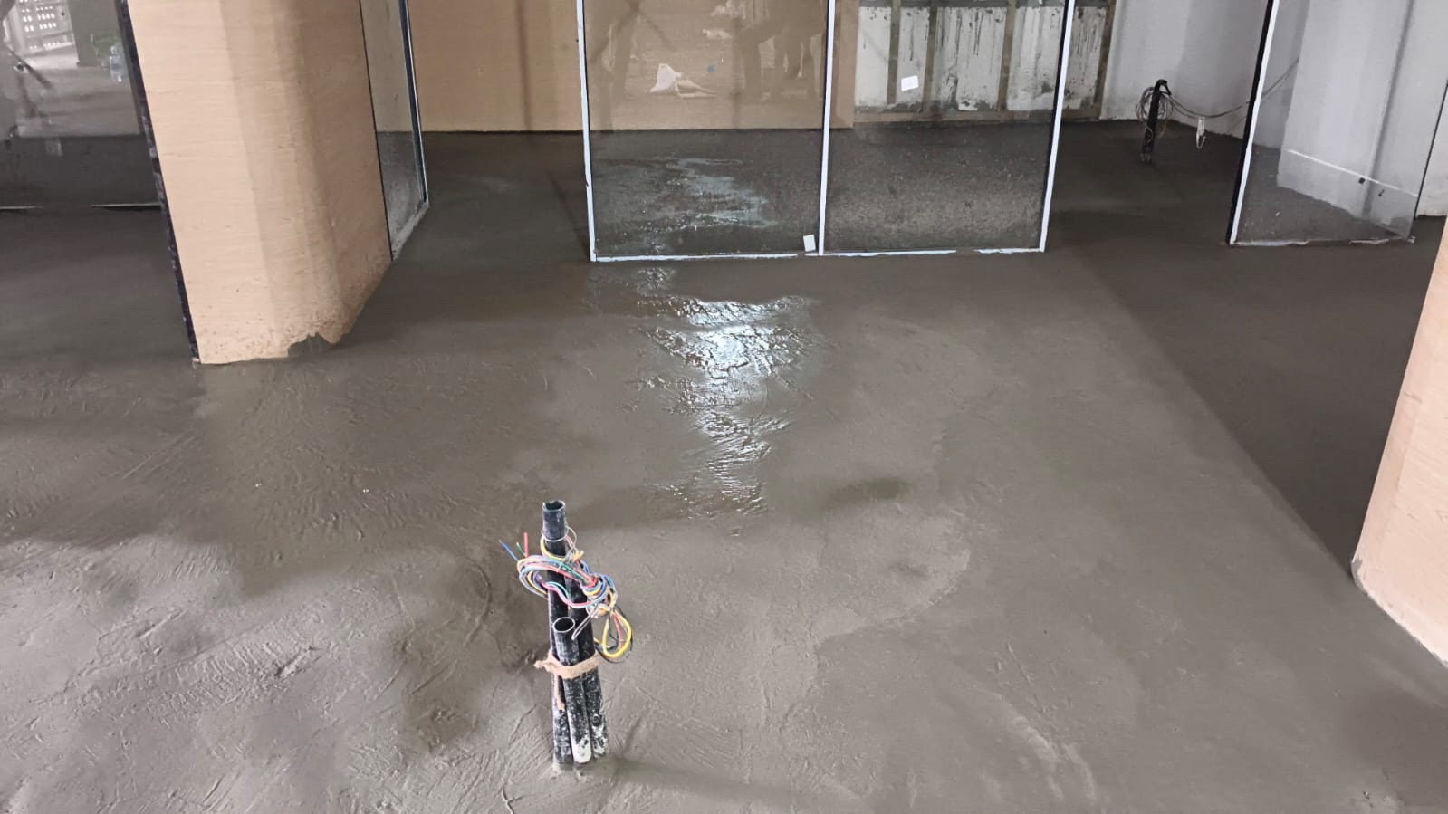 Site photograph, August 2025: fresh grey floor screed, still wet in places, with a bundle of conduits and coloured wires standing up through it.