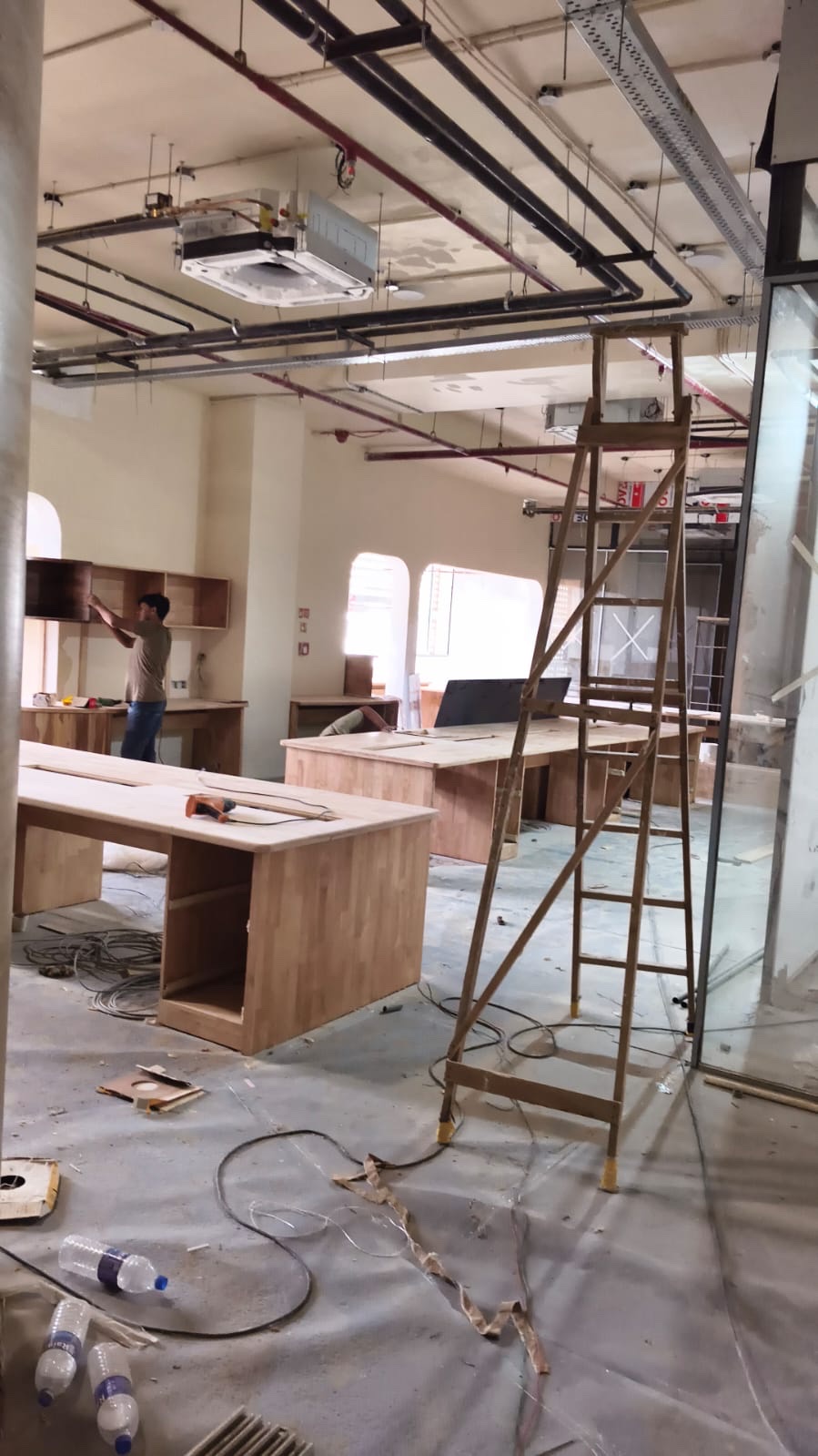 Site photograph, September 2025: wooden desks in place on a floor still covered with protective sheets, a tall ladder in the foreground, and someone fitting a wall cabinet at the back.