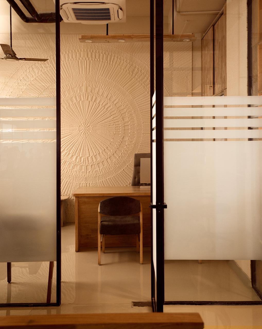 Seen through the cabin’s steel-framed glass front, a wall carved with a sunburst of radiating lines in pale plaster, with a desk and a chair in front of it.