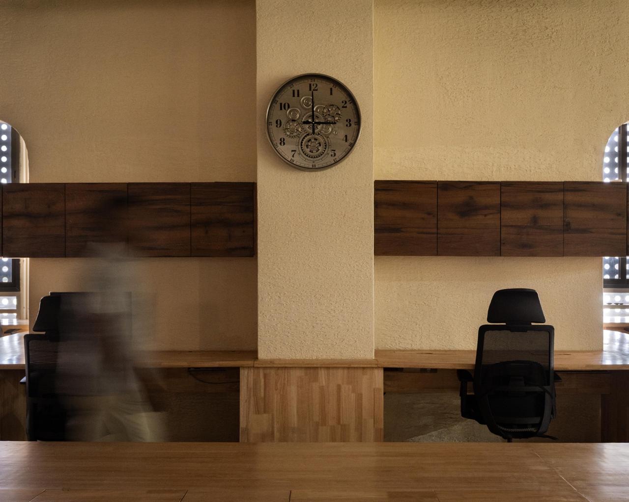 A run of workstations along a textured wall under dark timber cabinets, with a clock on the column between them. A blurred figure walks past on the left.